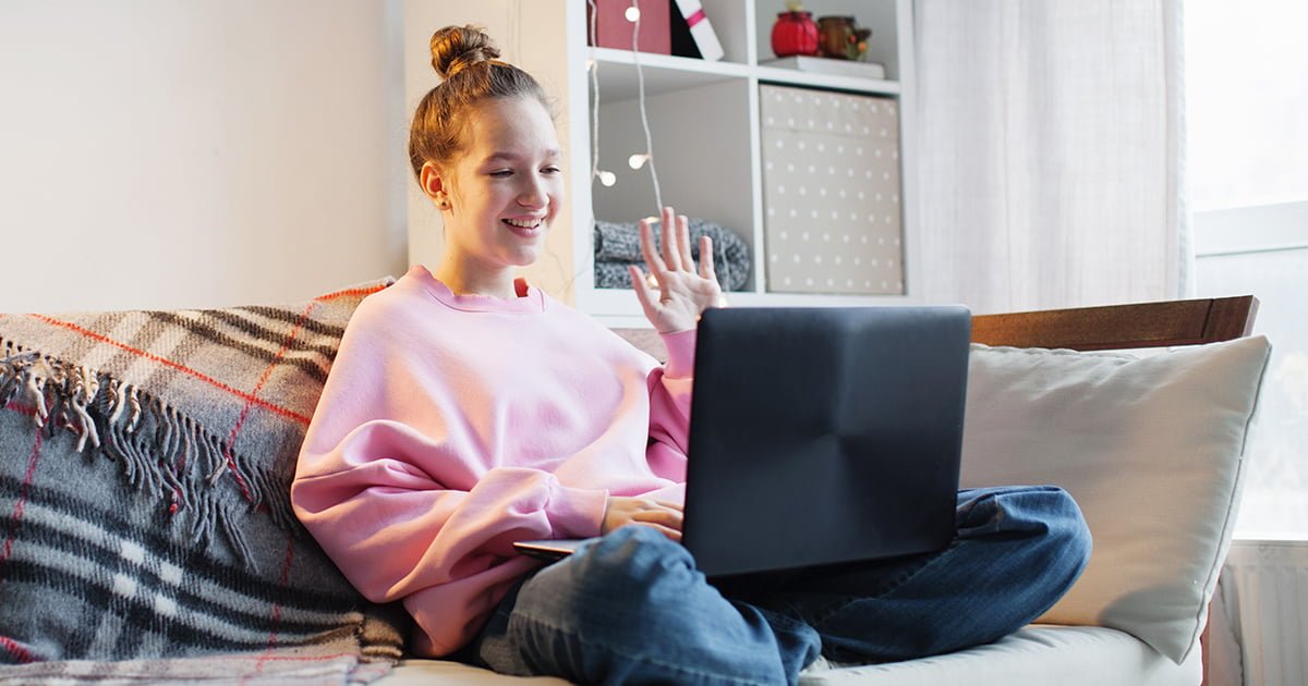 Tonåring sitter med en laptop i sitt knä och vinkar till någon på skärmen.
